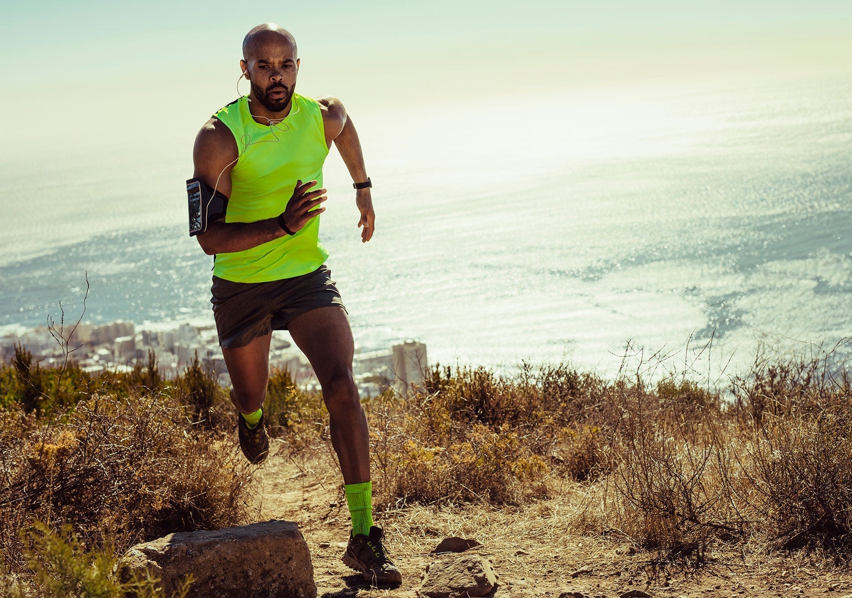 A man in a yellow shirt runs uphill, wearing earphones and an armband, on a grassy trail under a clear sky.