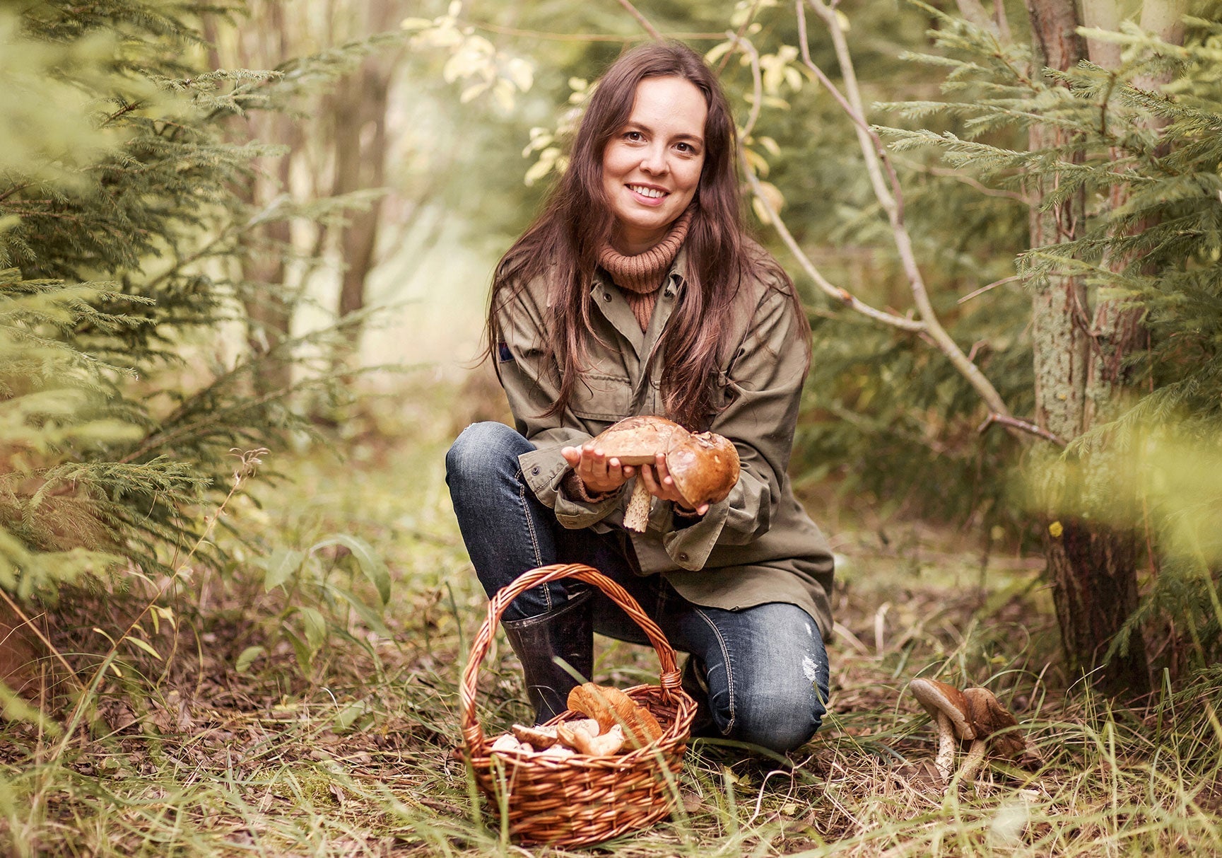 A woman squats in a forest, smiling and holding a basket full of various mushrooms, surrounded by trees and grass.