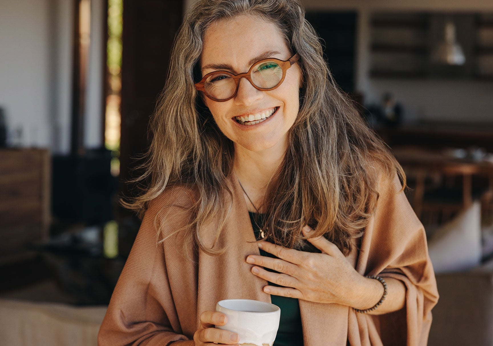 A woman wearing glasses smiles while holding a white coffee cup indoors. She wears a bracelet, and her hair is partially visible.