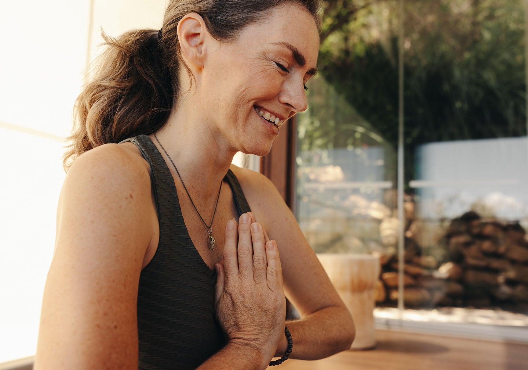 A smiling woman with clasped hands, wearing a necklace and bracelet, captured in an outdoor setting.