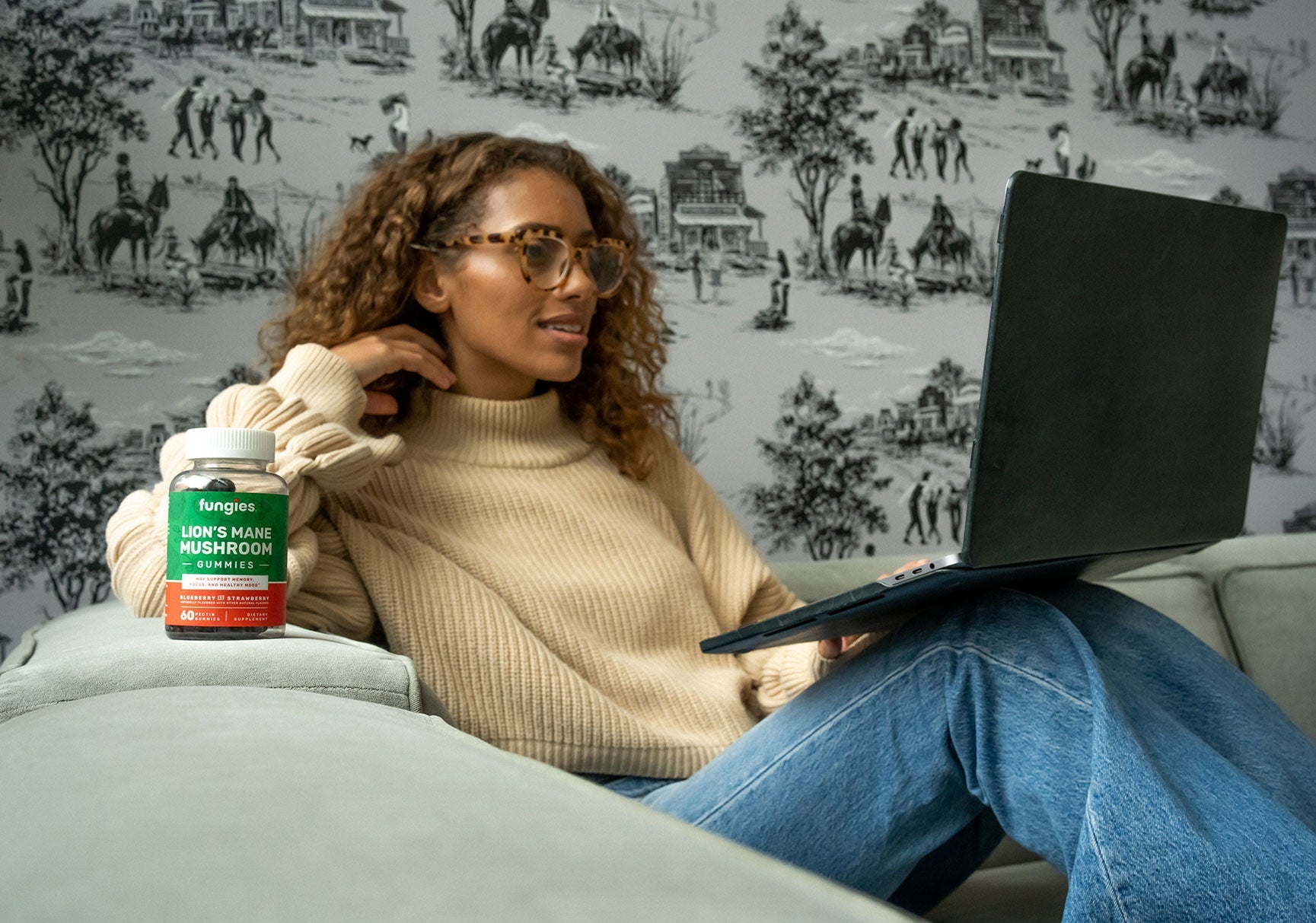 A woman wearing glasses sits on a couch using a laptop, with a bottle of gummy mushrooms visible beside her.