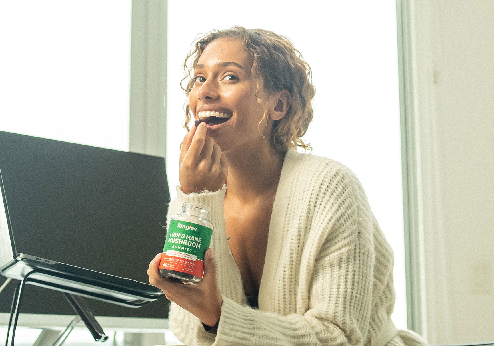 A woman indoors eating a cookie, with a bottle and a can of juice visible nearby, while using a laptop.