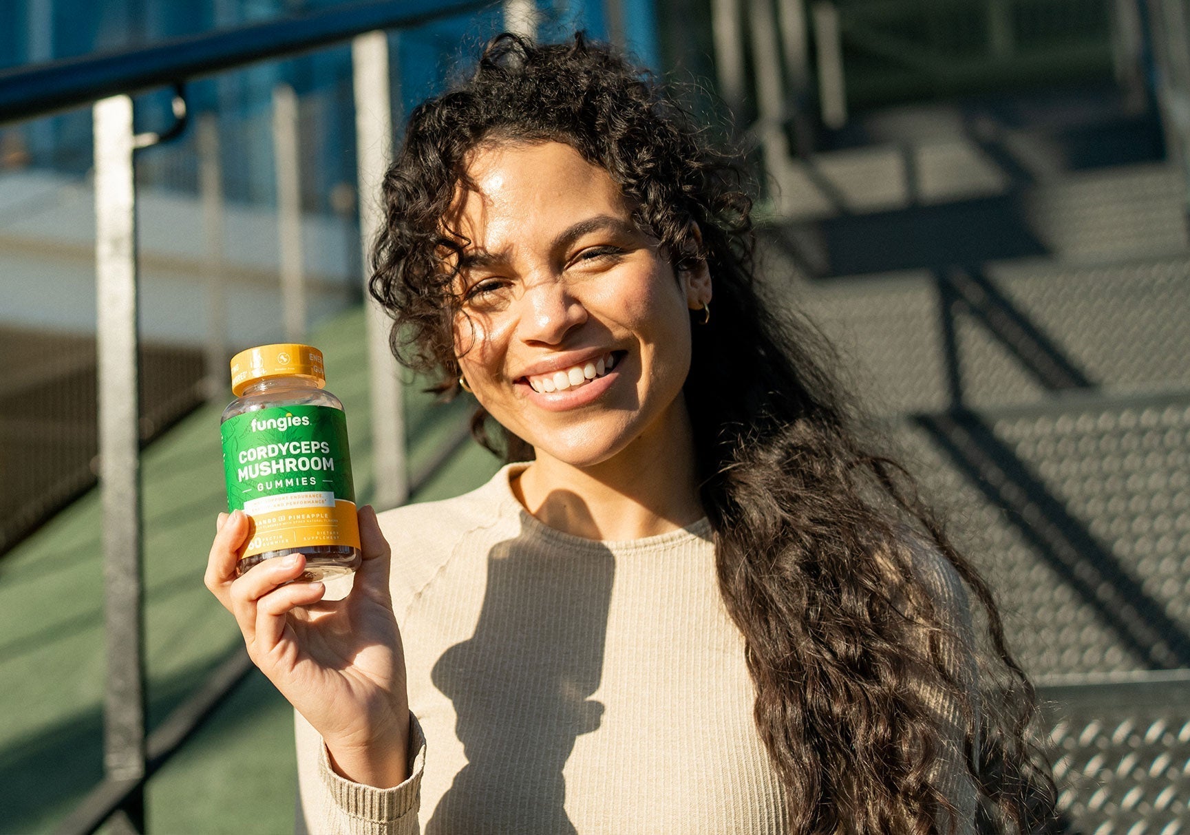 A woman smiling holds a bottle of vitamins, with her hand visible. The image highlights her face and the product she is showcasing.