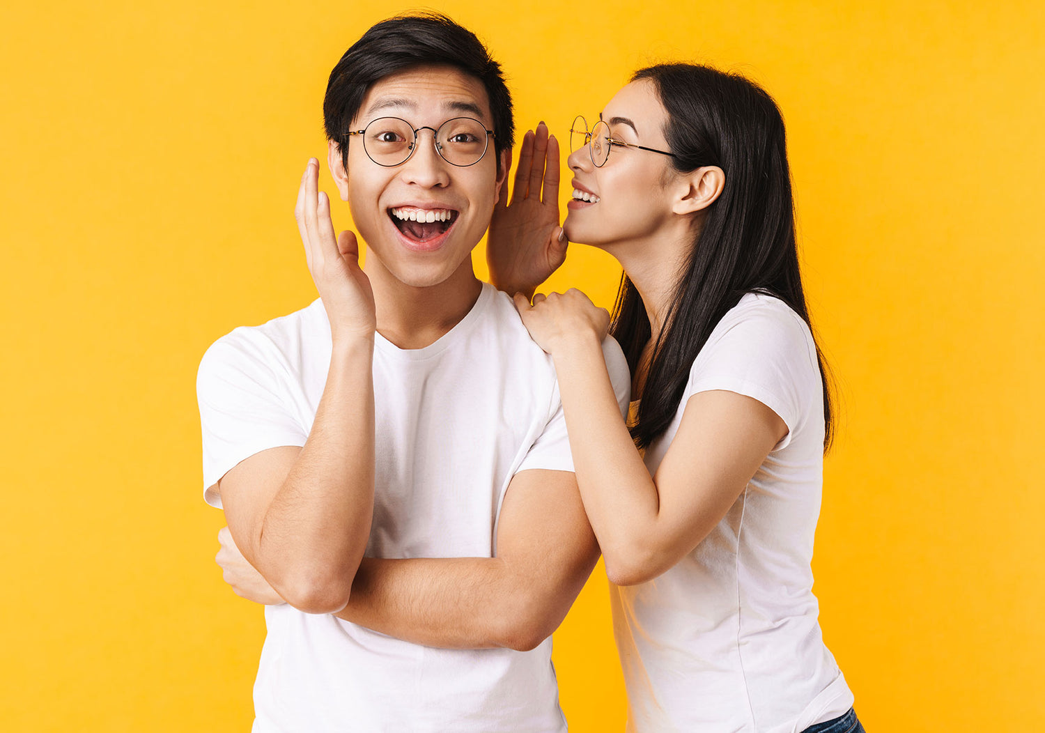 Image of smiling woman in eyeglasses whispering secret to surprised asian man isolated over yellow background