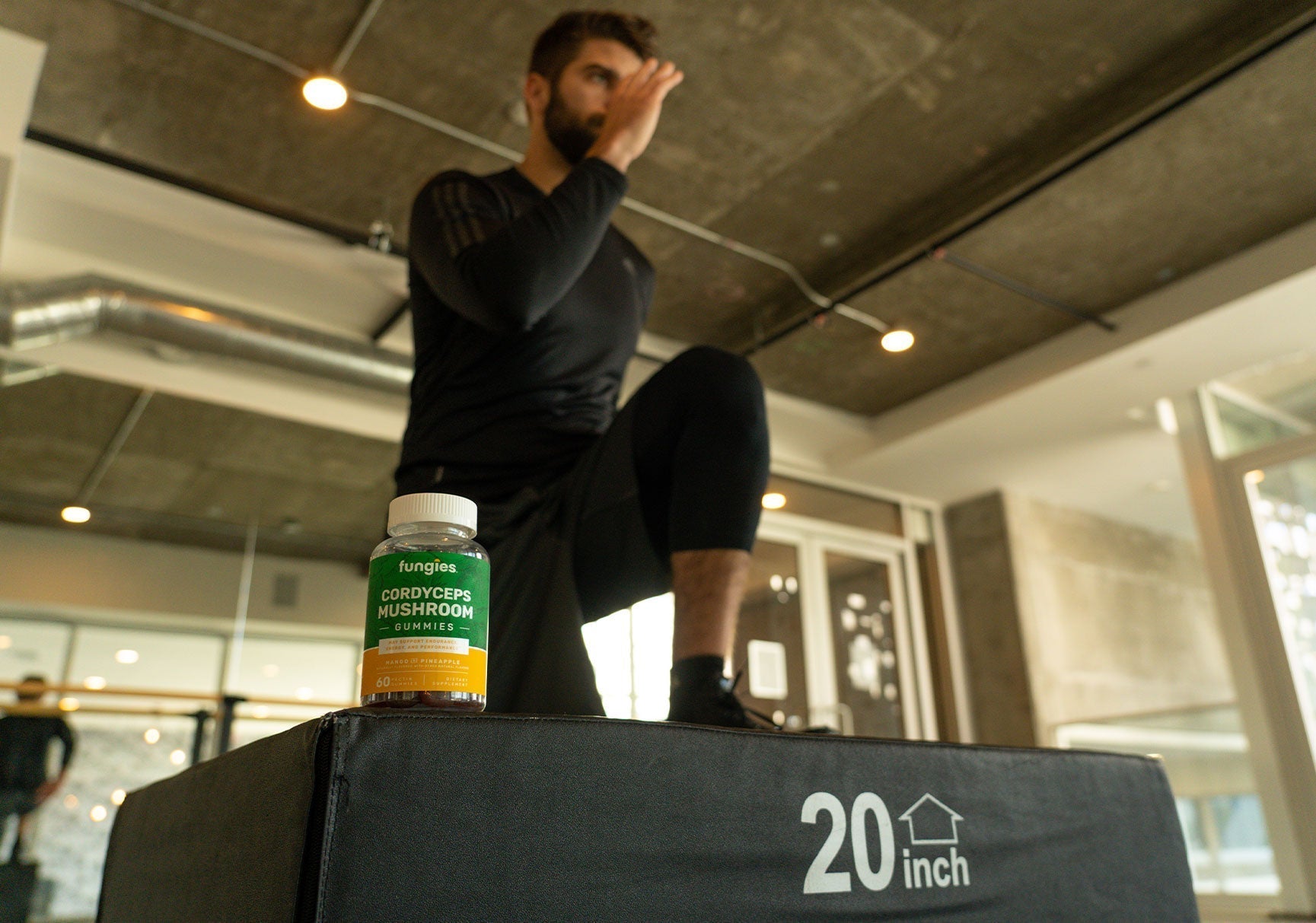 A man in a black shirt squatting on a box indoors, next to a close-up of a bottle, with a dimly lit background.