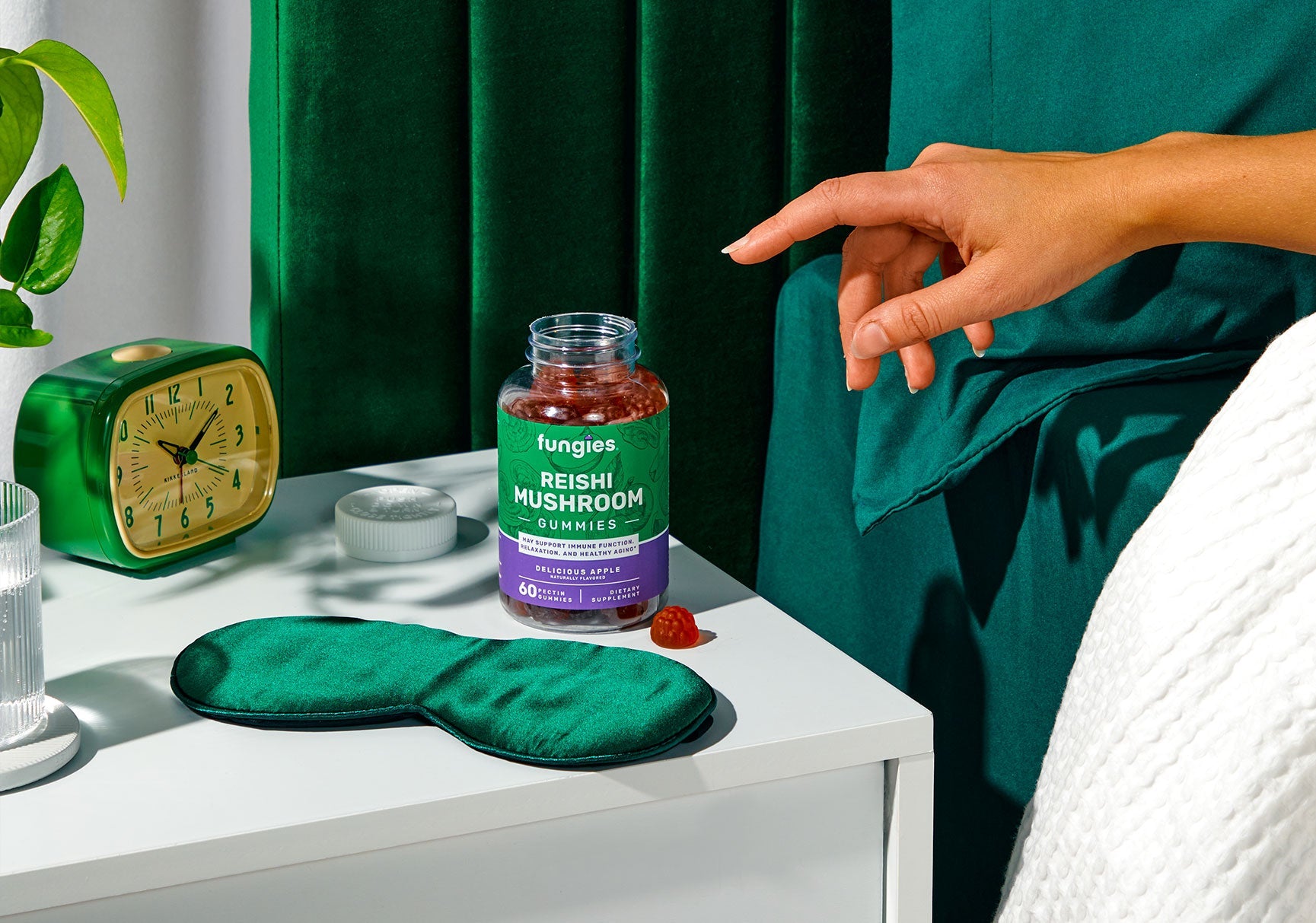 A hand points at a jar of mushroom jam, alongside a green eye mask and a jar of gummy candies on a table.