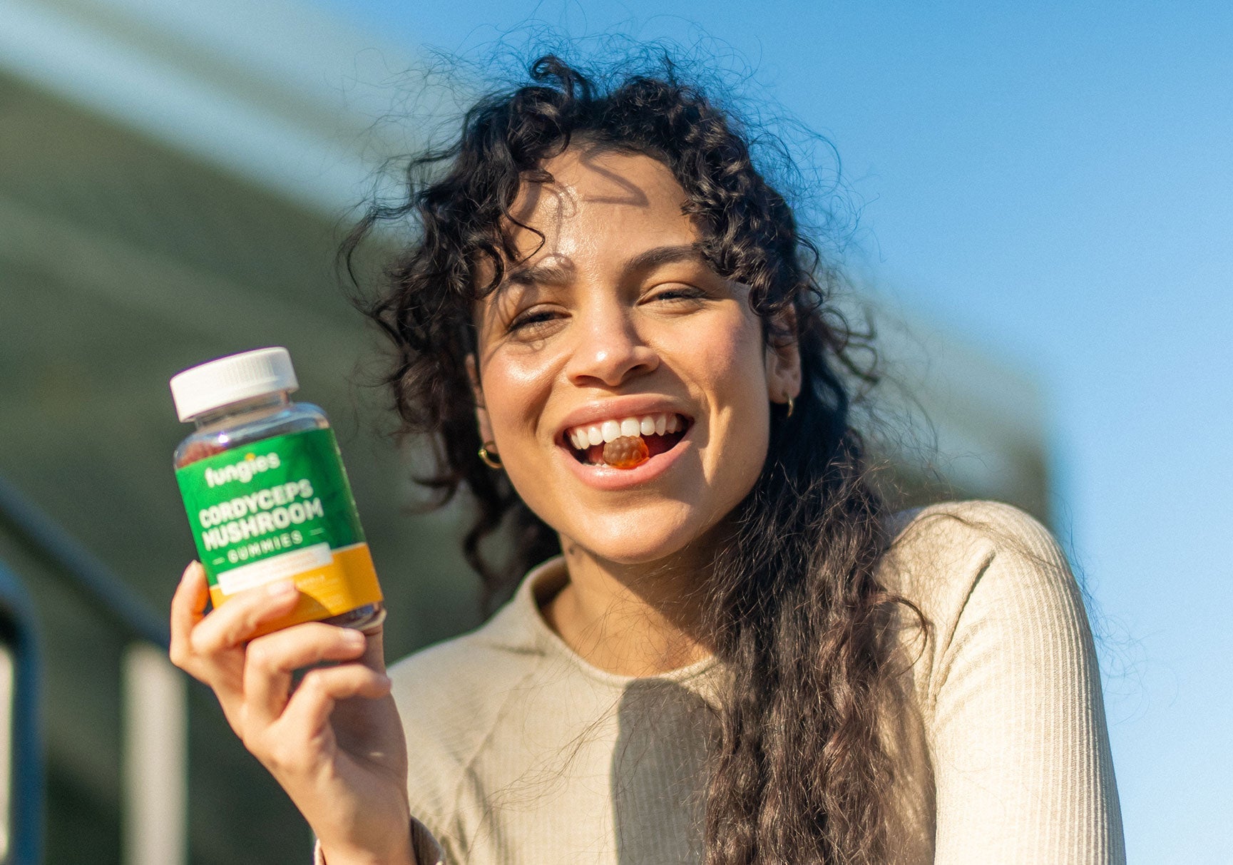 A woman smiling while holding a vitamin bottle, with a close-up of her mouth containing a gummy candy.