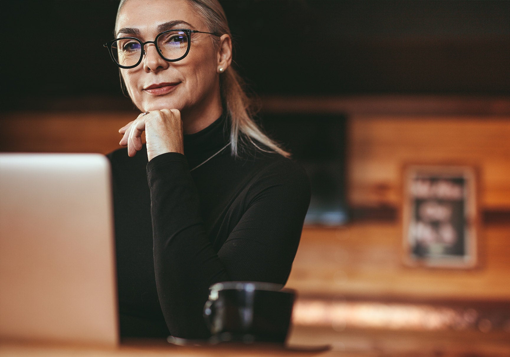 A woman in glasses and a black turtleneck sits at a table, resting her chin on her hand, with a laptop nearby.