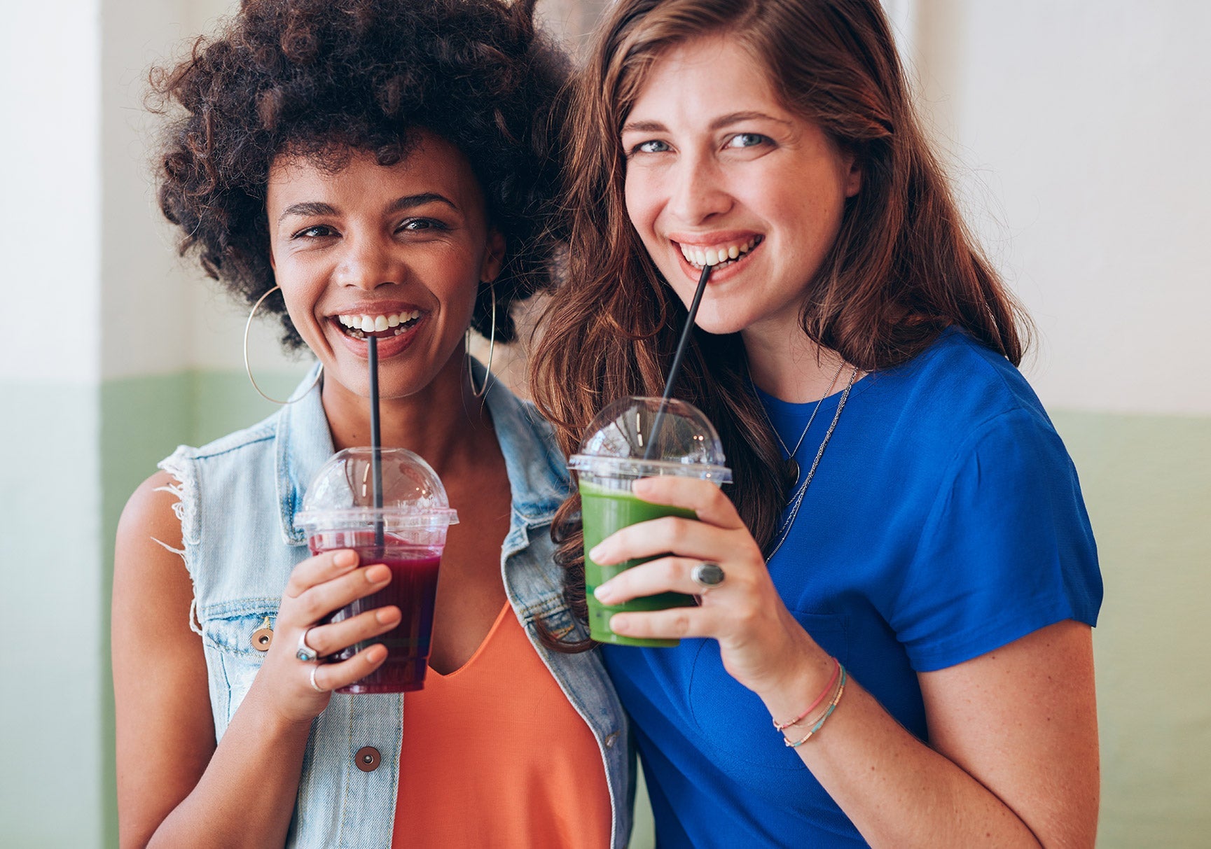 Two women smiling and holding green drinks with straws indoors, showcasing casual enjoyment and friendship.