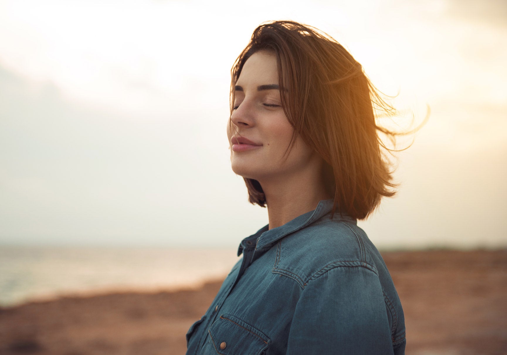 A woman with long brown hair, eyes closed, stands on a beach, wearing a blue shirt, captured in an outdoor portrait.