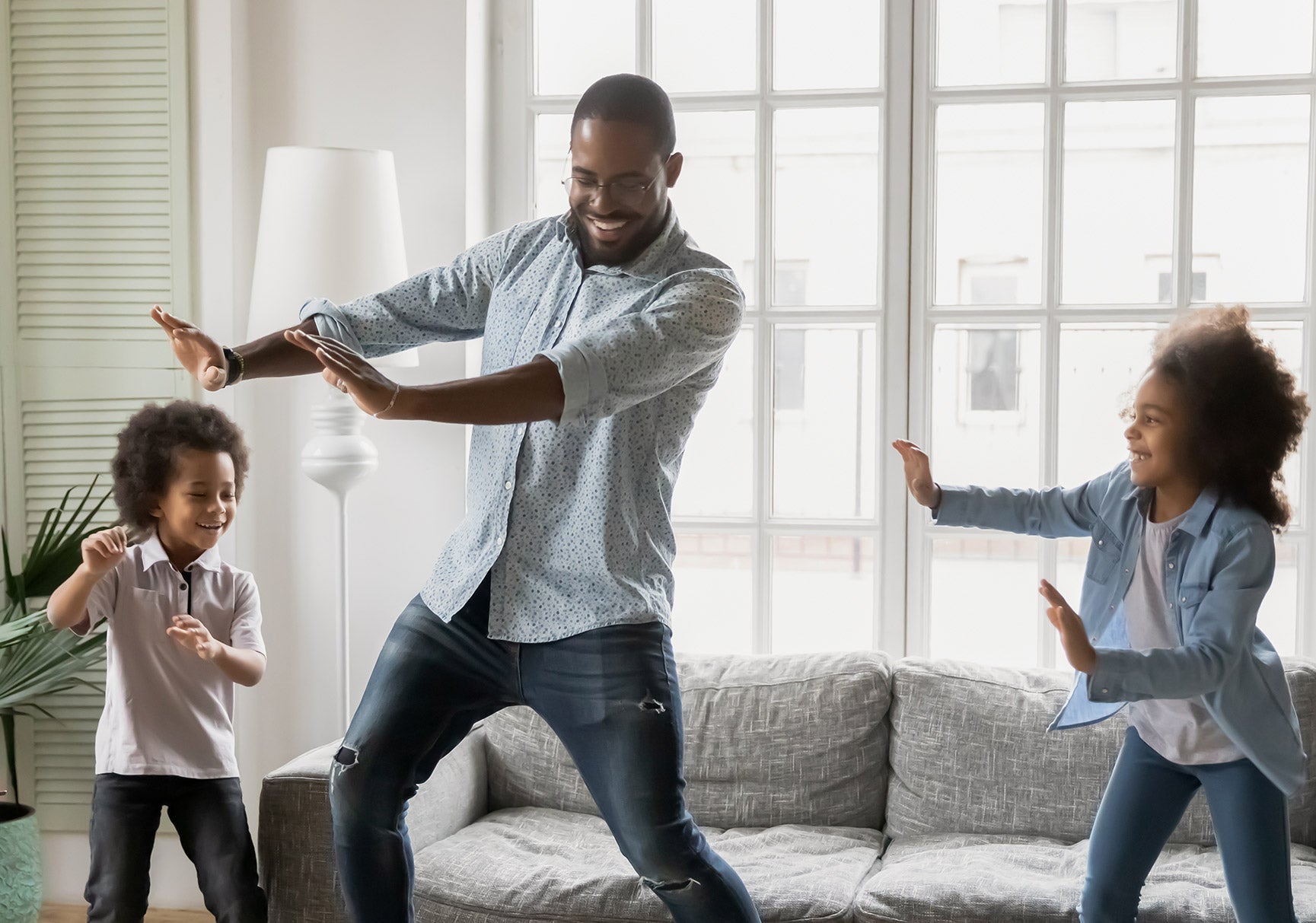 A man and two children dance joyfully in a living room, with one child holding a brush and another standing on a couch.