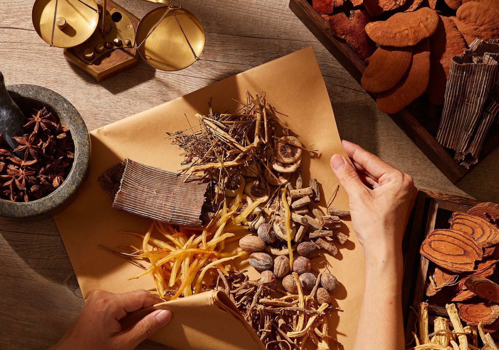 A person wraps paper around various foods, including a bowl of star anise and a box of mushrooms, with a scale nearby.