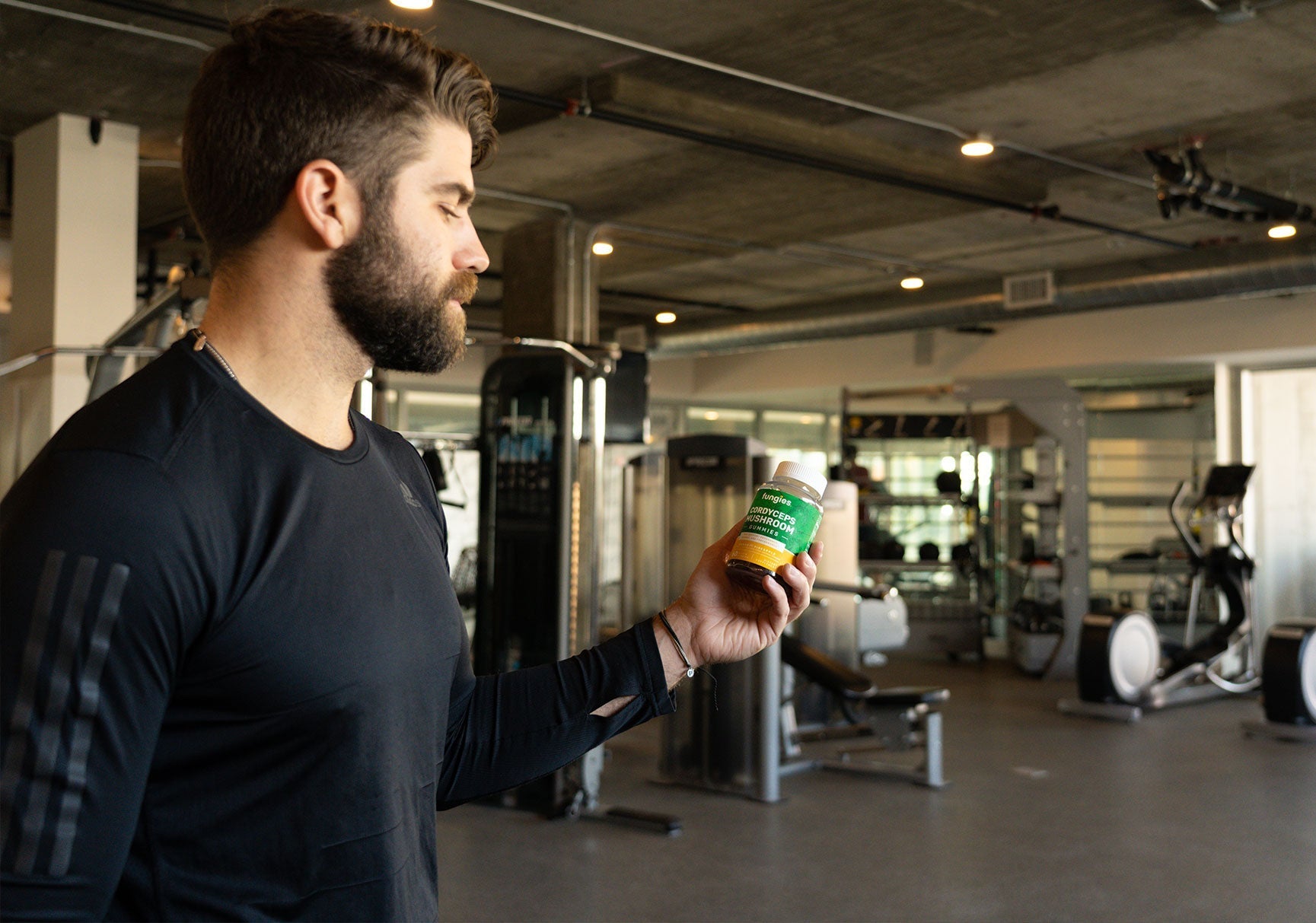 A bearded man in a black shirt holds a bottle of vitamins, standing indoors next to gym equipment.