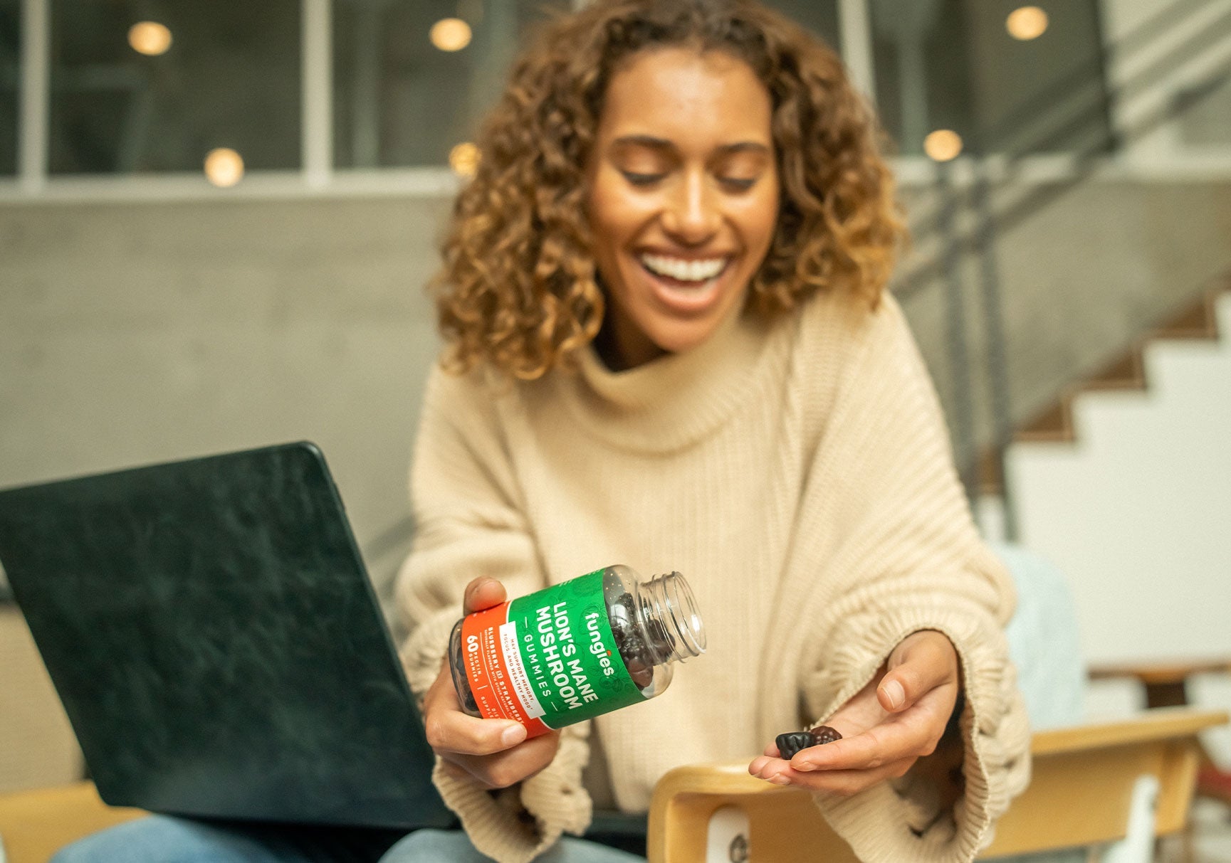 A smiling woman with curly hair holds a bottle of mushroom supplement, sitting indoors with a laptop in the background.