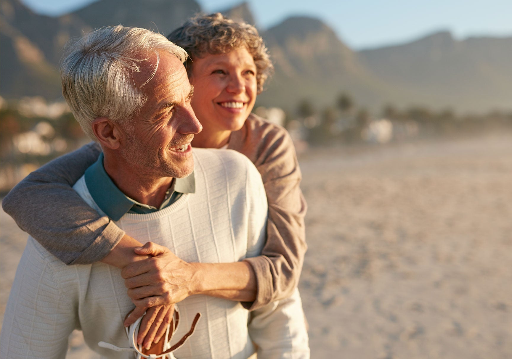 A man carries a woman on his back outdoors, both smiling. The background suggests a beach setting with blurred elements.