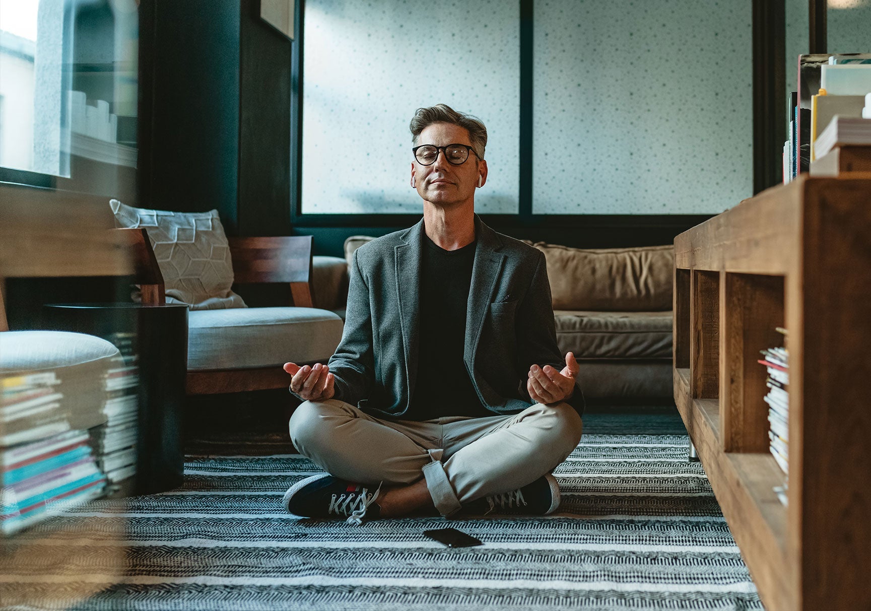 A man with glasses and earbuds sits cross-legged on the floor in a room with bookshelves and scattered books.