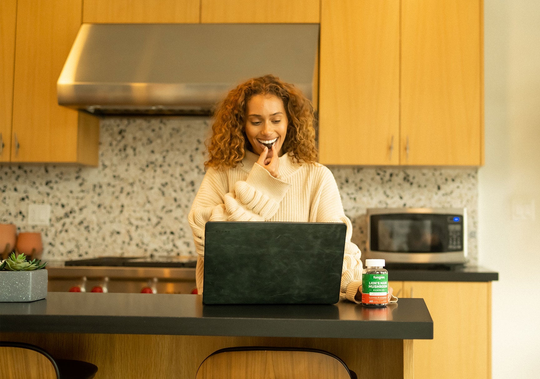 A woman in a white sweater sits at a table with a laptop, eating a cookie. A bottle of mushroom vitamins is nearby.