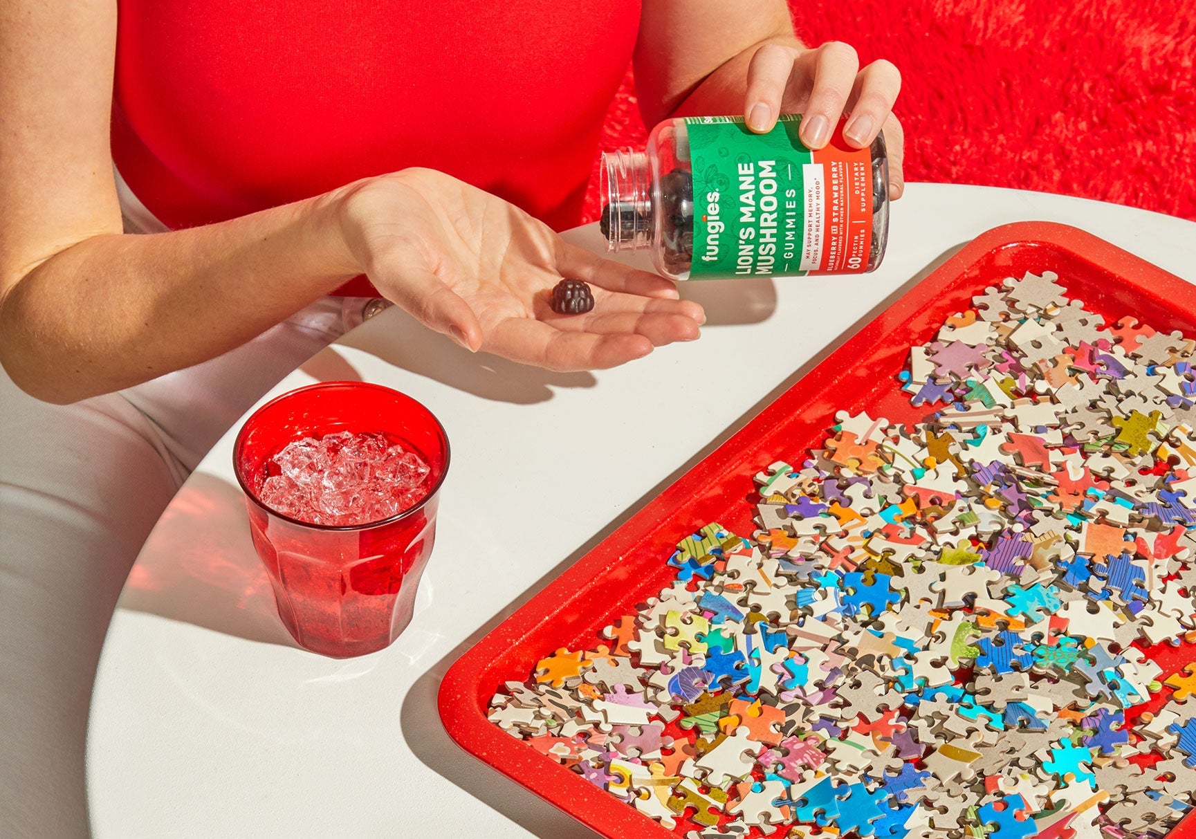 A person holding a jar of gummy mushrooms near a red glass filled with ice, suggesting a combination of supplements and refreshment.