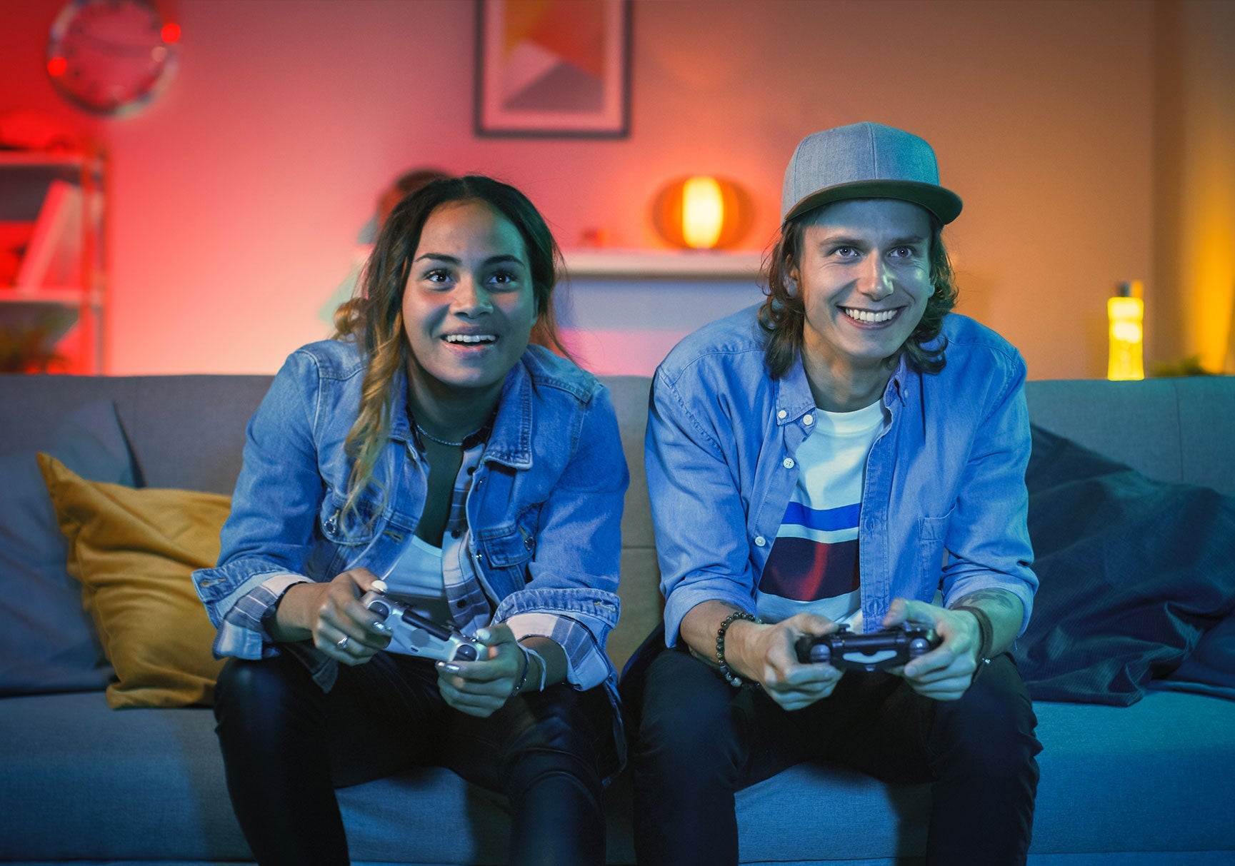 A man and woman sitting on a couch, smiling, playing video games with controllers in hand, in a cozy indoor setting.