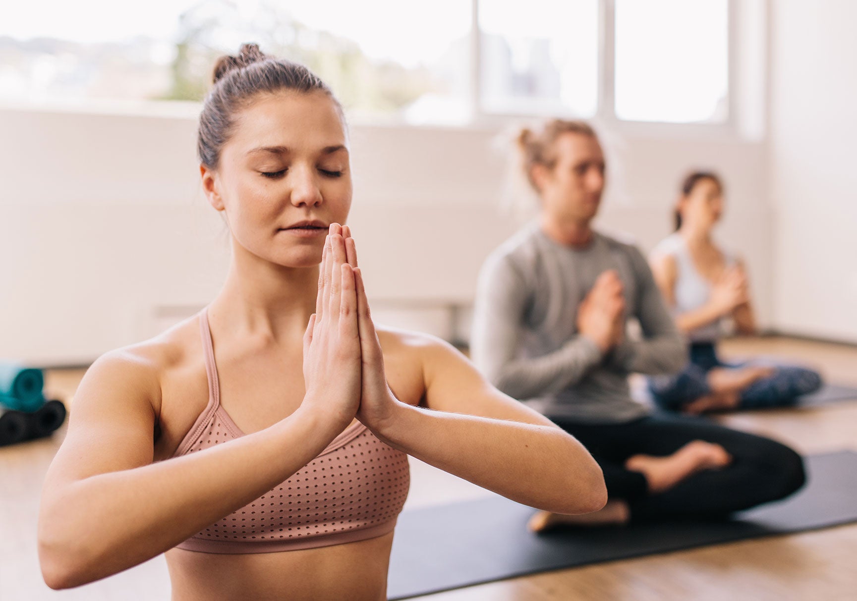 A woman practicing yoga indoors, posed with eyes closed and hands together, alongside a group meditating in the background.