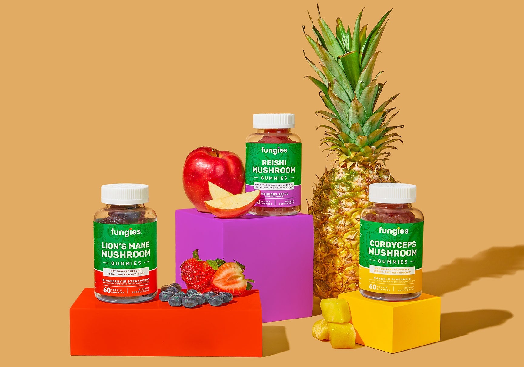 Bottles of vitamins and jars of gummy mushrooms surrounded by fresh fruit, including a pineapple, blueberries, and apples, displayed on a table.