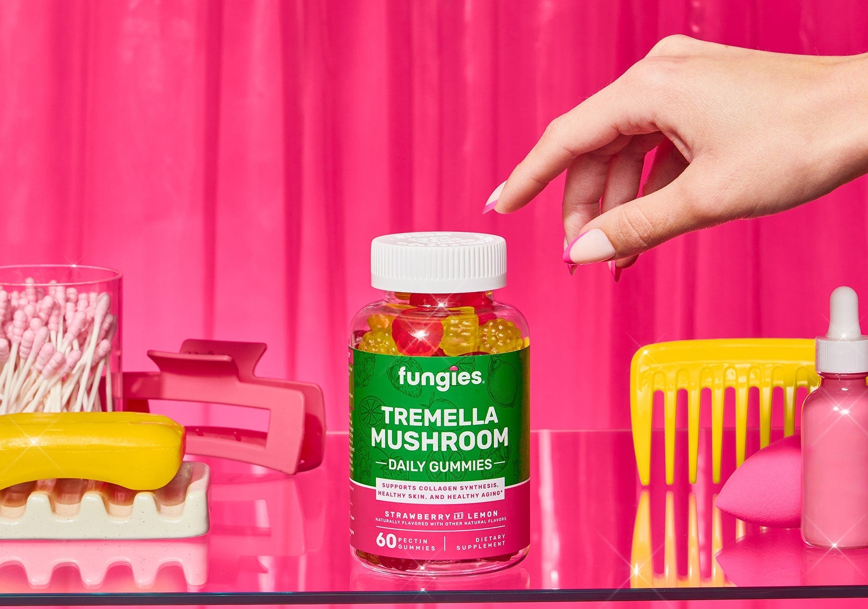 Hand reaching for a jar of gummy candies with a pink nail polish bottle nearby, set against a simple indoor background.